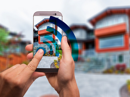 Hände halten ein Smartphone. Dieses steuert Geräte eines Smart Home an.