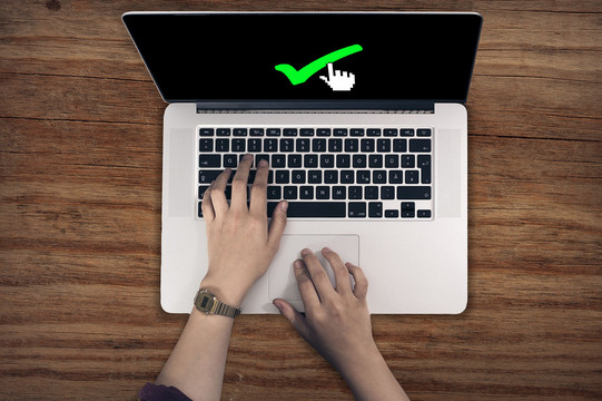 Eine Person tippt mit der linken Hand auf der Tastatur eines Laptops und fährt mit der rechten Hand über das Touchpad. Auf dem Bildschirm ist der Mauszeiger auf einen grünen Haken gerichtet.