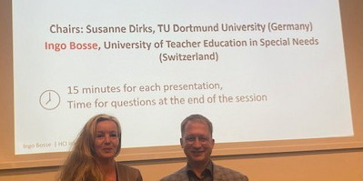 Auf der linken Seite steht Dr. Susanne Dirks und rechts daneben Prof. Dr. Ingo Bosse. Im Hintergrund sieht man die Eröffnungsfolie für die Session.